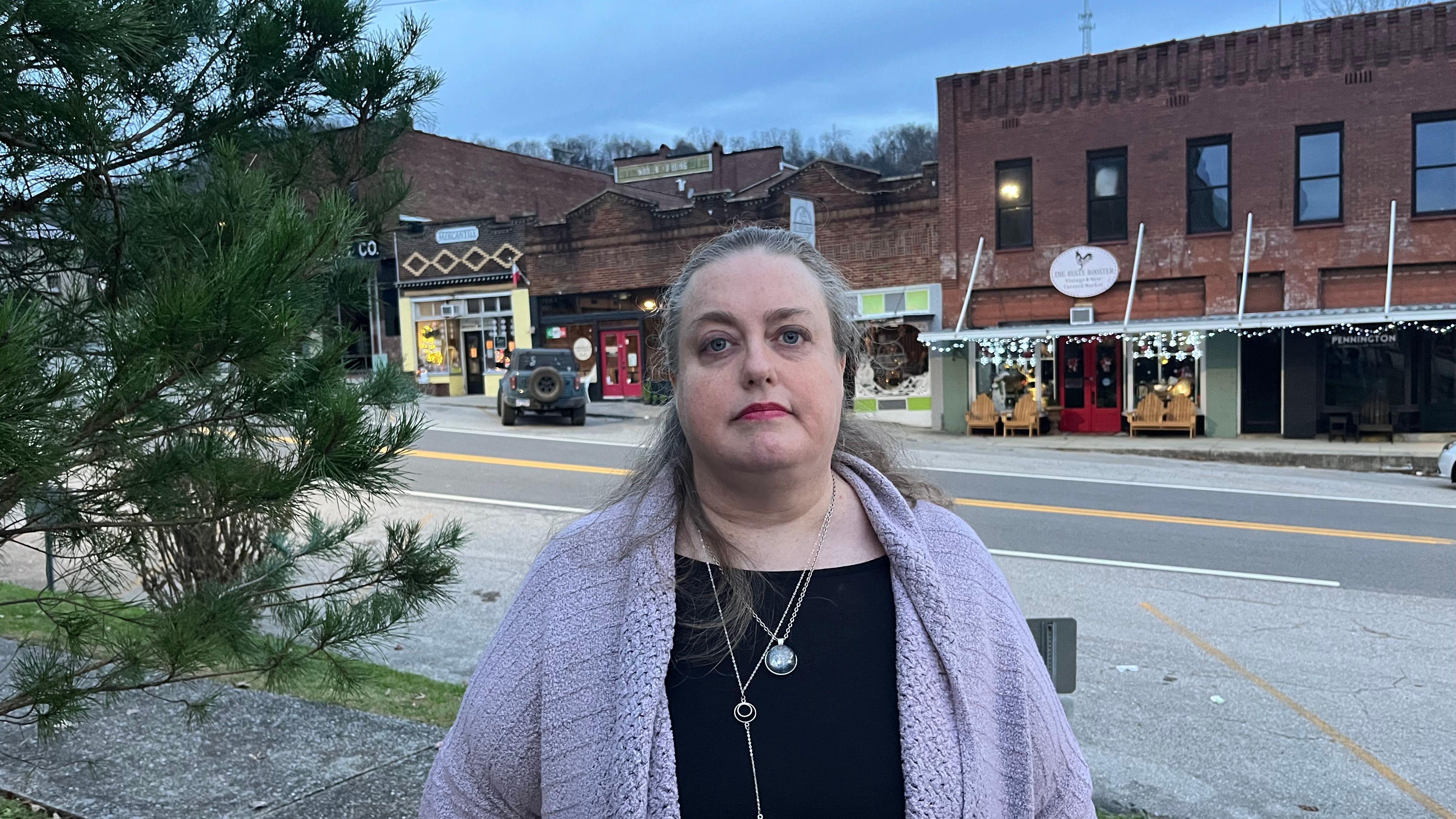 Mulher com cabelo grisalho e casaco lil&aacute;s, de p&eacute; em frente a uma s&eacute;rie de lojas, na pra&ccedil;a principal de Gainesboro.