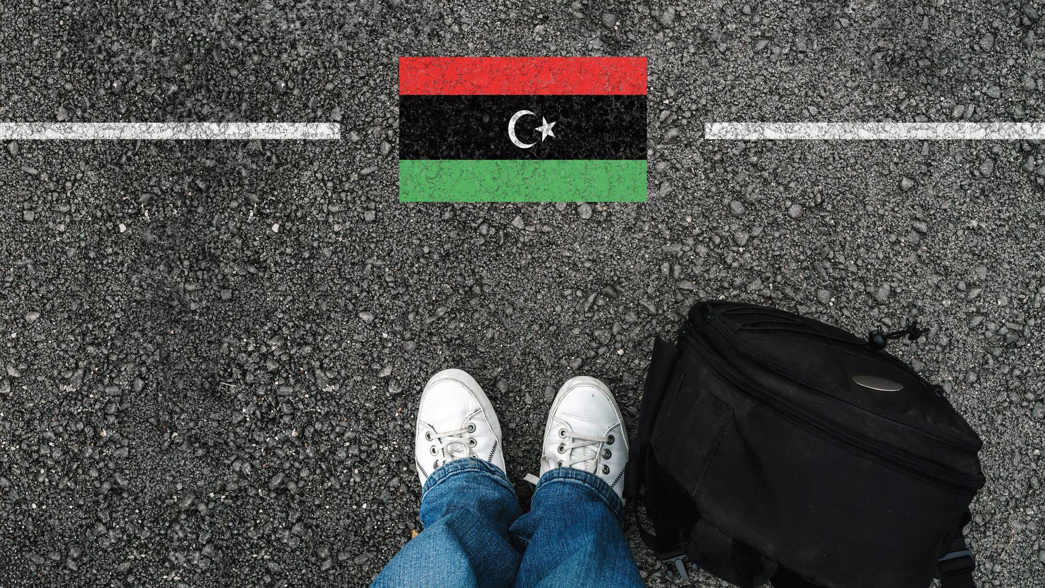 الحكومة الليبية تنفي اتفاقاً مع واشنطن بشأن ترحيل مهاجرين، وتؤكد رفضها التوطين القسري على أراضيها 3 رجل يرتدي حذاءً وحقيبة ظهر يقف على الأسفلت بجوار علم ليبيا والحدود.