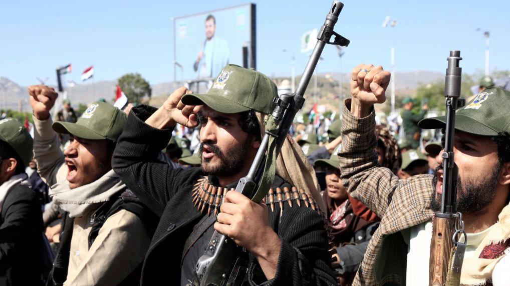 مقاتلو جماعة الحوثي في صنعاء: ثلاثة رجال يحملون بنادق ويرفعون قبضاتهم وخلفهم صورة لعبد الملك الحوثي