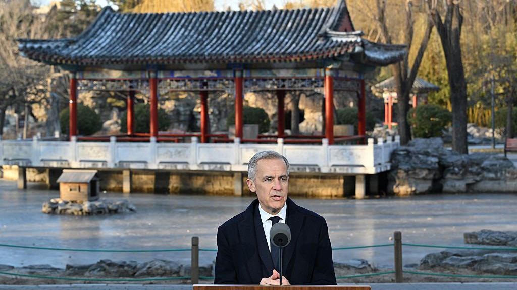 El primer ministro canadiense, Mark Carney, de traje y corbata, habla a un micrófono. Tras él, una construción tradicional china.