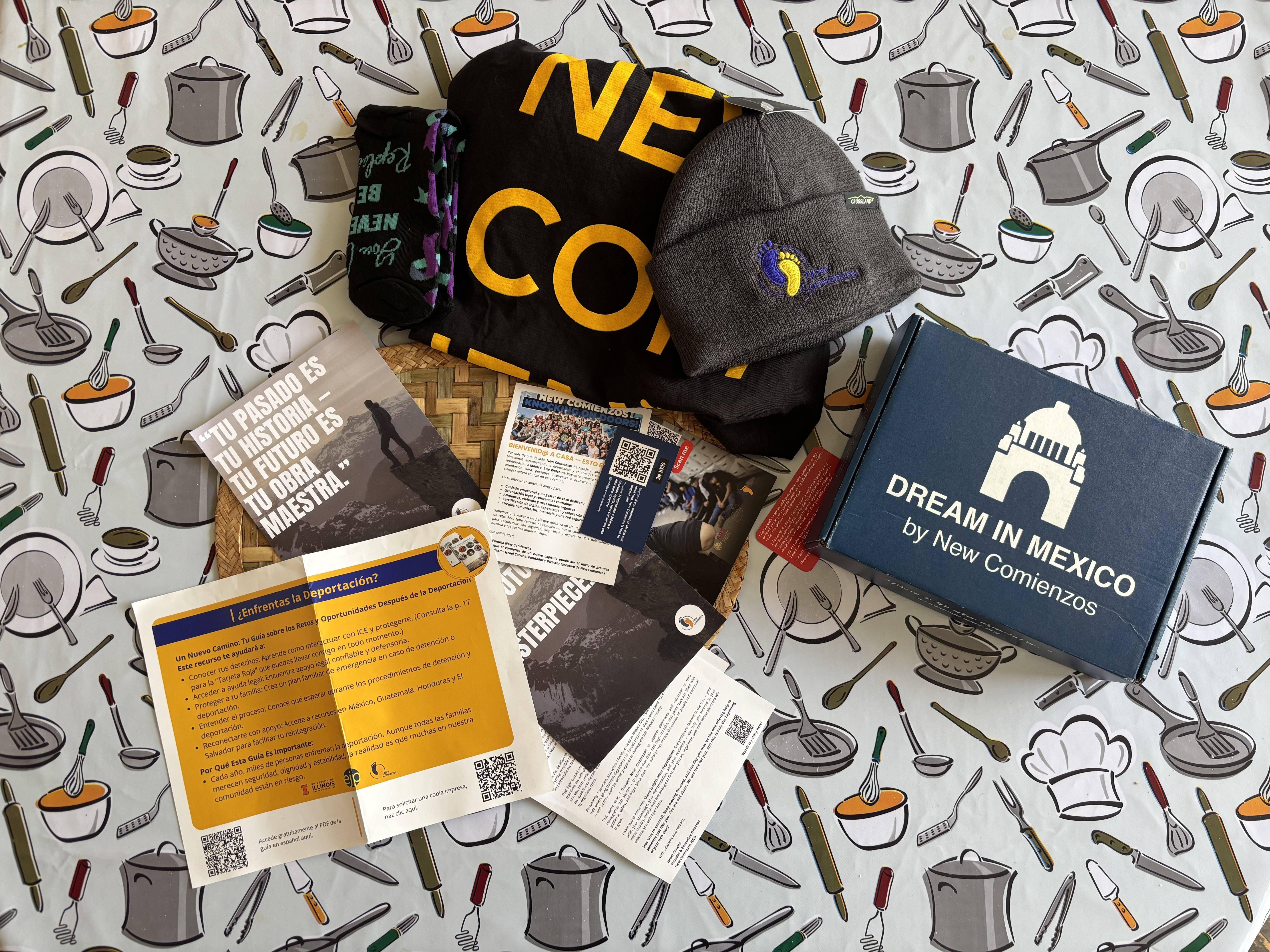 Artículos promocionales y folletos de New Comienzos, incluidos gorros, una bufanda y un libro, colocados sobre un mantel con ilustraciones de utensilios de cocina.