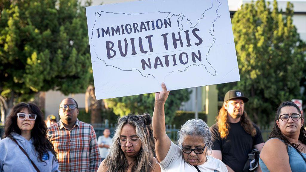Manifestantes protestam contra uma deportação realizada pelo ICE em Los Angeles, na Califórnia (EUA). Uma manifestante, com a cabeça baixa, exibe um cartaz com os dizeres 'A imigração construiu esta nação'.