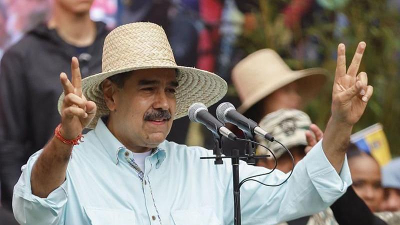 مادورو يرتدي قميصاً يميل إلى اللون الأزرق ويرتدي قبعة من القش مخاطباً الجماهير ورافعاً علامة النصر بكلتا يديه.