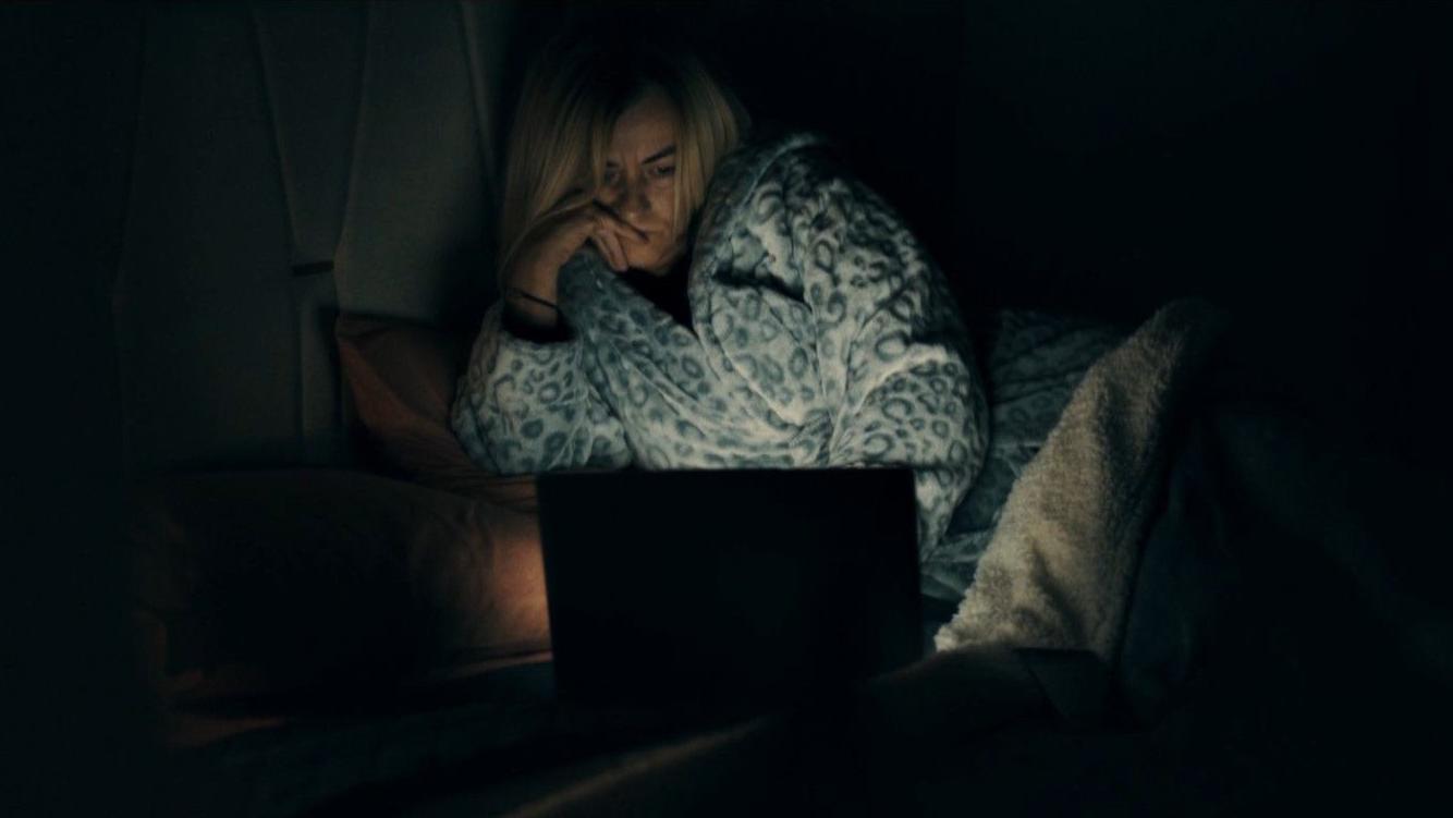 Uma mulher de pijama tem o rosto iluminado por um laptop; ela apoia as m&atilde;os no rosto, pensativa.