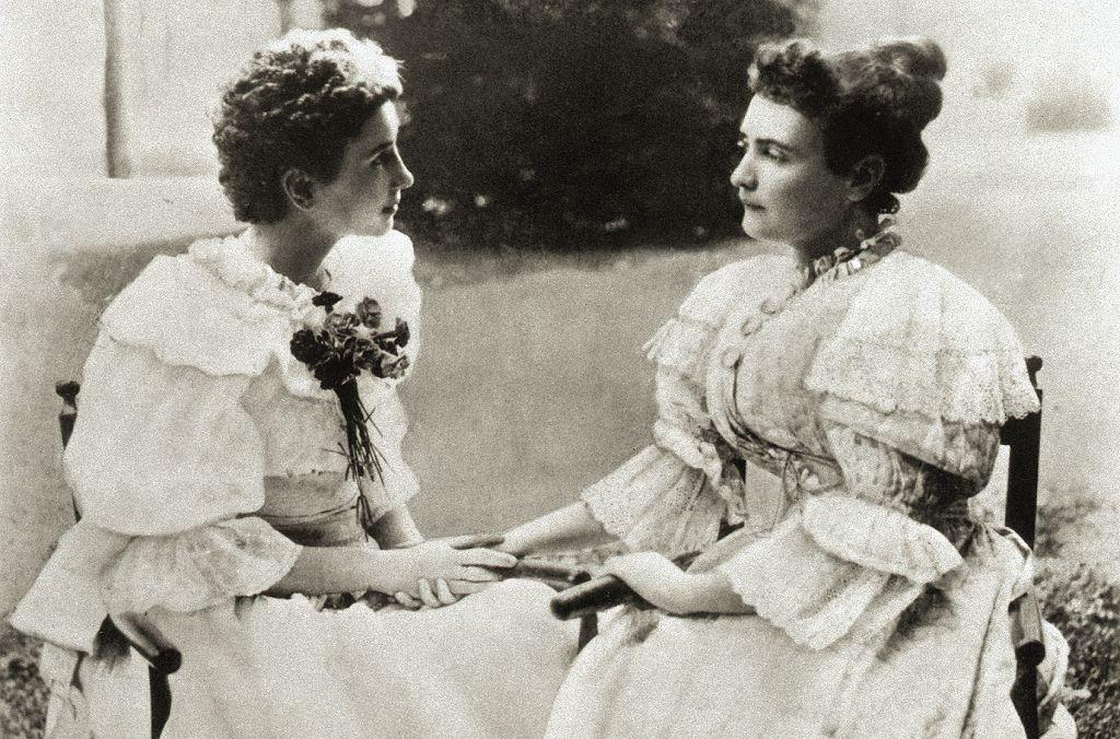 Las dos mujeres, sentadas en un jardín, con vestidos claros de mangas embombadas, con encajes y lazos. Se miran a los ojos y tienen sus manos juntas, pues era su forma de comunicación. 