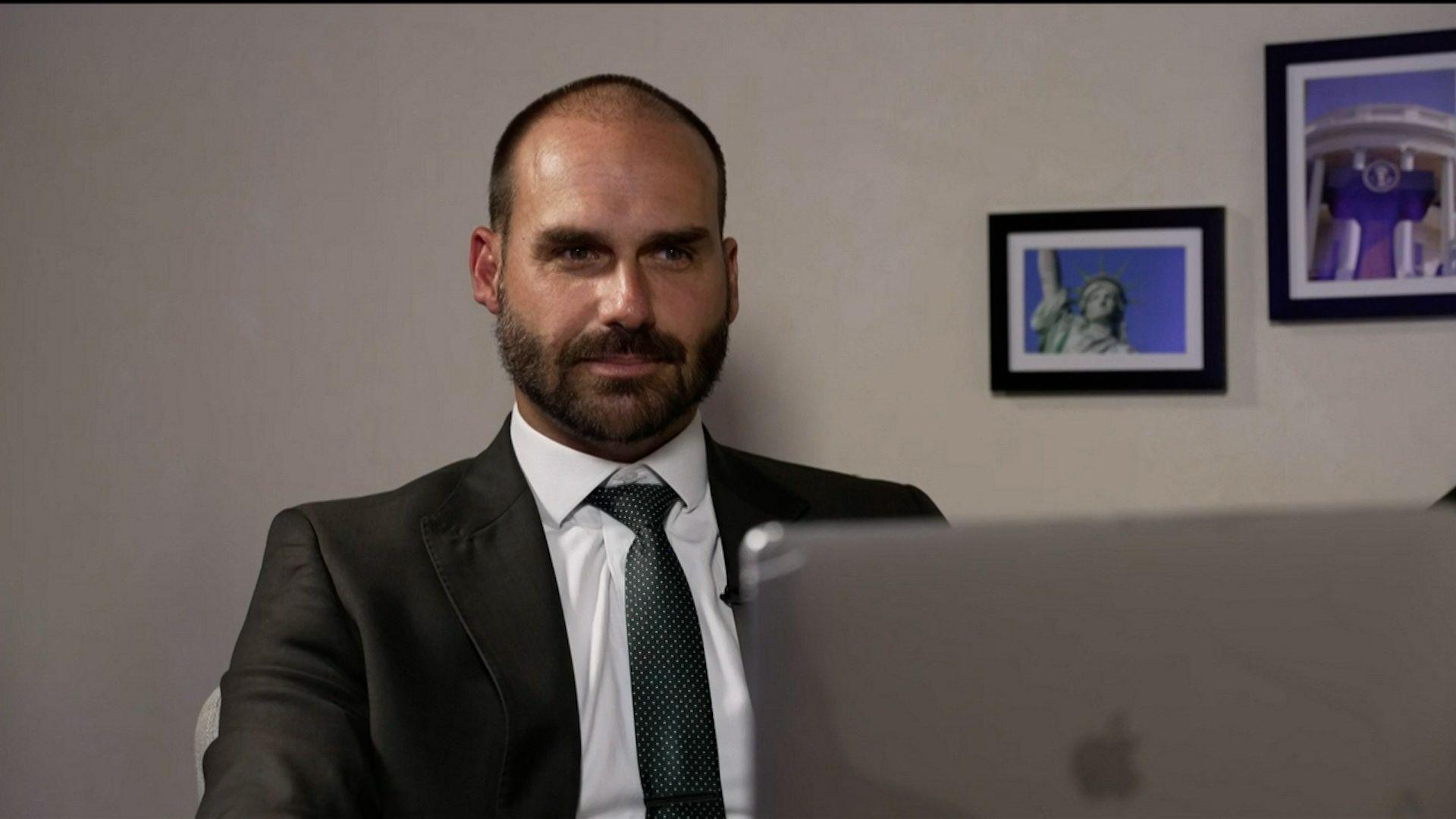 Eduardo Bolsonaro sorrido de forma contida e olhando para laptop