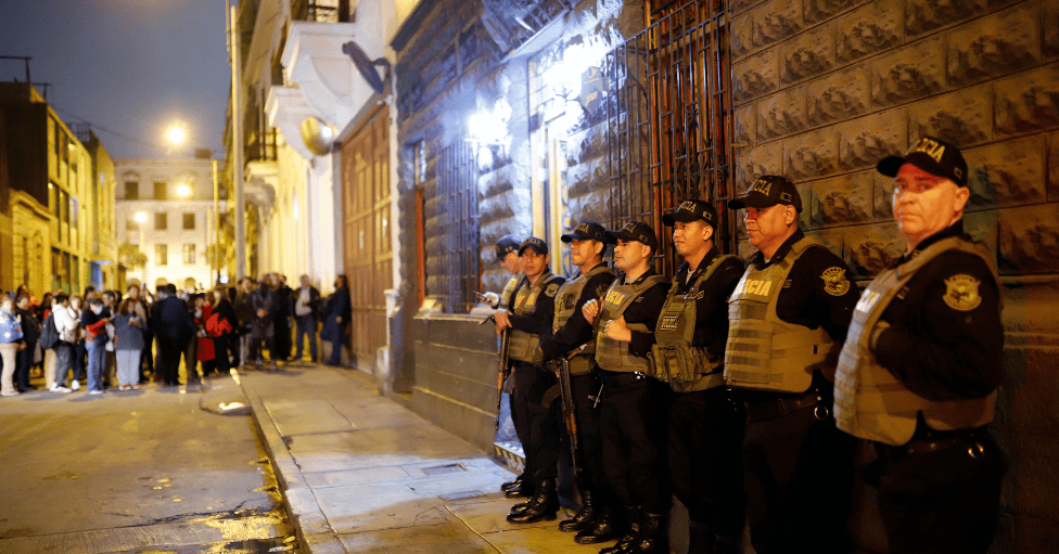 Un grupo de policías, de pie junto a un edificio. Es de noche y al fondo, a unos metros, en la misma calle, se distingue a un grupo de personas. 