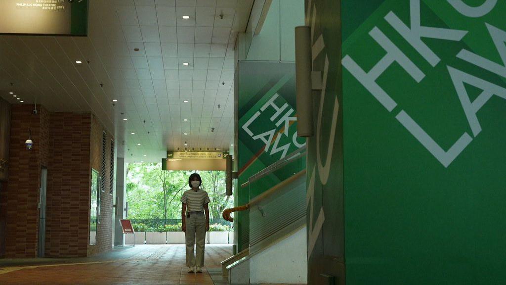 A young woman (Siu Kwong) stands in an empty corridor next to a staircase flanked by green pillars with HKU LAW written in white capitals. 