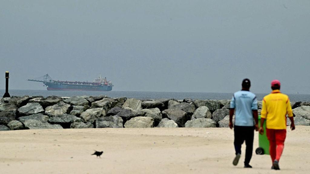 Dos personas de espaldas caminando por una playa bajo el sol. A lo lejos, en el mar, un buque de carga. 