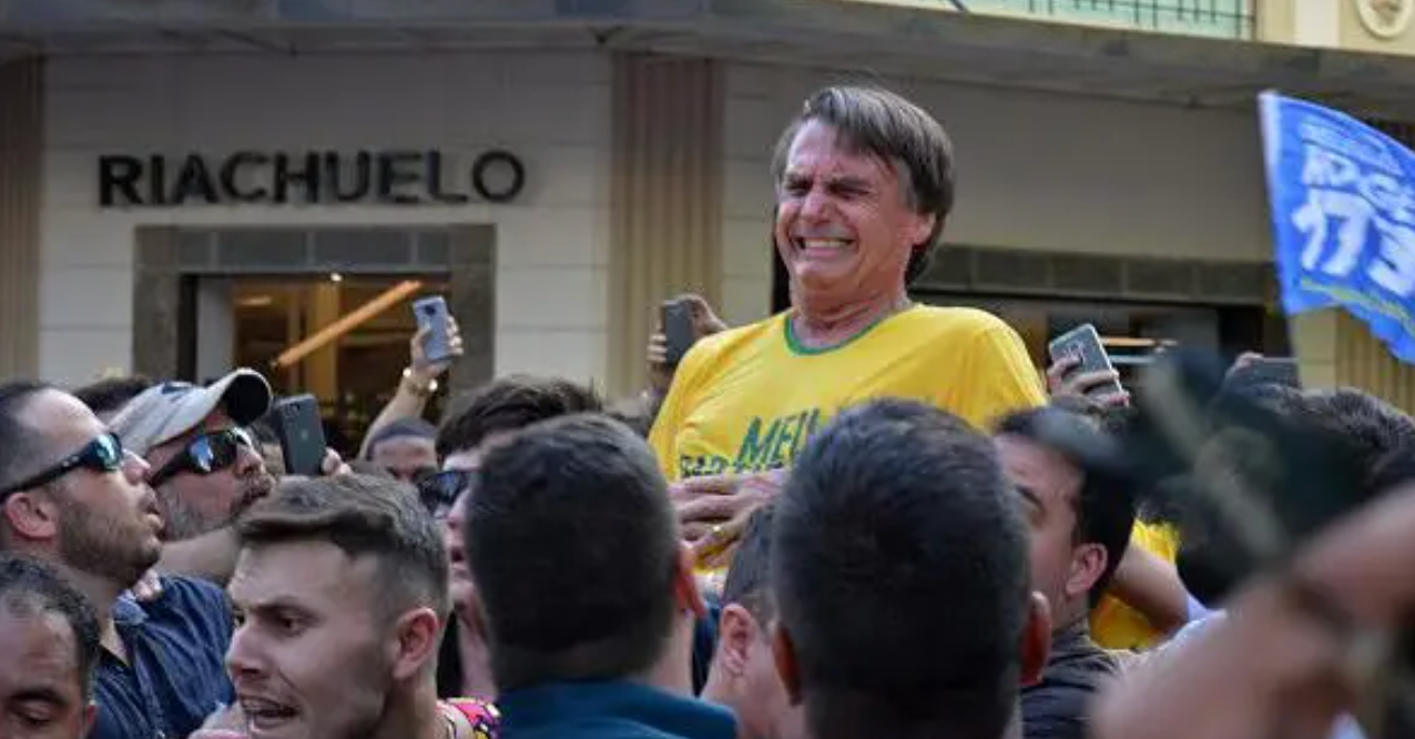 Foto mostra Bolsonaro no momento em que recebeu facada. Est&aacute; usando uma camisa amarela e fazendo uma cara de dor em meio &agrave; multid&atilde;o