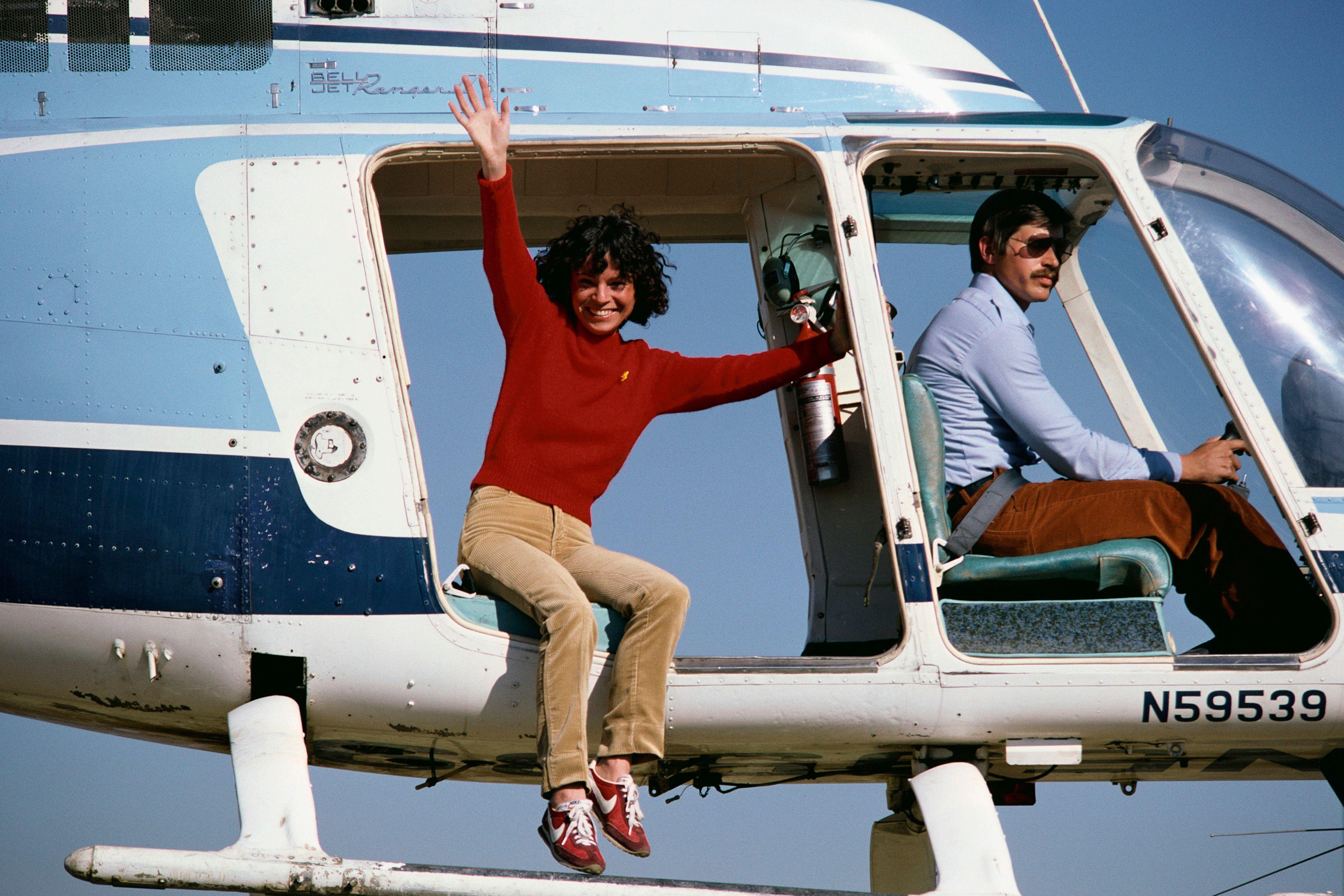 Kitty O'Neil con un suéter rojo y pantalones marrones sentada en el borde de un helicóptero en el aire, con los pies colgando. Ella está sonriendo y saludando. Un piloto masculino con gafas de sol y bigote está volando el avión. Es un día soleado y brillante.