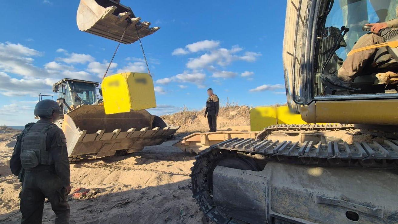 Las tropas israelíes observan cómo una excavadora coloca un bloque amarillo en su sitio. El trabajo cuenta con el apoyo de otro vehículo de construcción, y el bloque queda suspendido en el aire mediante una cadena fijada a la excavadora. 