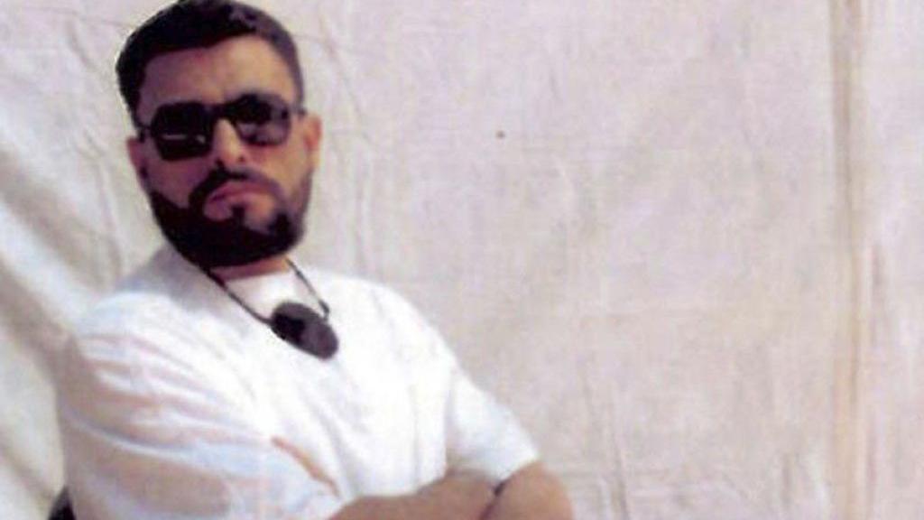 أبوزبيدة يرتدي نظارة شمس سوداء وتي شيرت أبيض وبنطلون أبيض ويجلس على كرسي