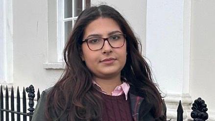 Maryam Khan de p&eacute;, no lado externo de uma constru&ccedil;&atilde;o com uma grade de ferro. De &oacute;culos, camisa rosa com colarinho, blusa vinho e jaqueta verde, ela sorri levemente para a c&acirc;mera.