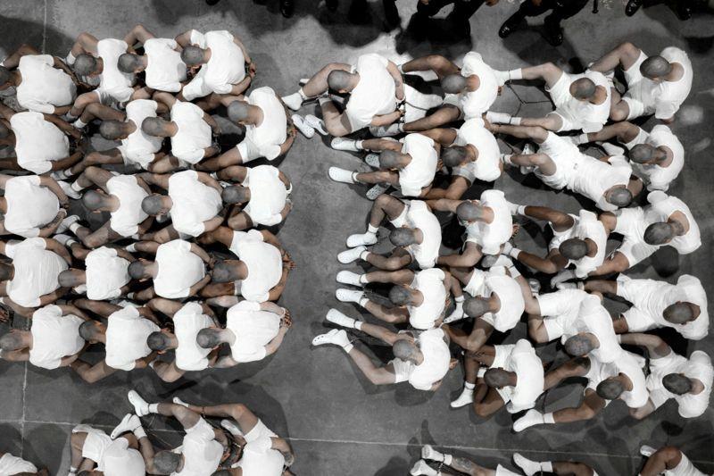 Filas de presos sentados no chão, com a cabeça abaixada e as mãos para trás
