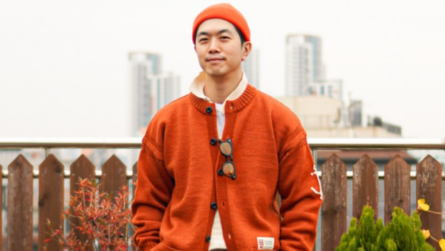 Ji Seung-ryeol sorri para a c&acirc;mera, com as m&atilde;os nos bolsos. Ele veste um su&eacute;ter e um gorro laranja.