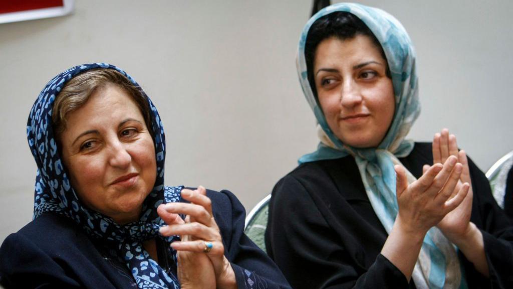 Shirin Ebadi y Narges Mohammadi aplauden durante una conferencia de prensa. Ebadi lleva un pañuelo estampado en azul oscuro, con algo de cabello visible, mientras que Mohammadi lleva un pañuelo estampado en azul claro, con parte de su cabello visible, y una chaqueta negra.