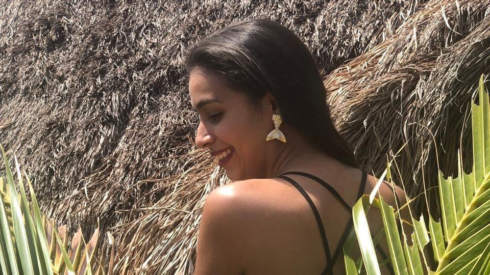 Catalina, entre ramas de palma, mira hacia un lado y sonríe mientras su espalda da a la cámara. En el fondo, un techo de paja.