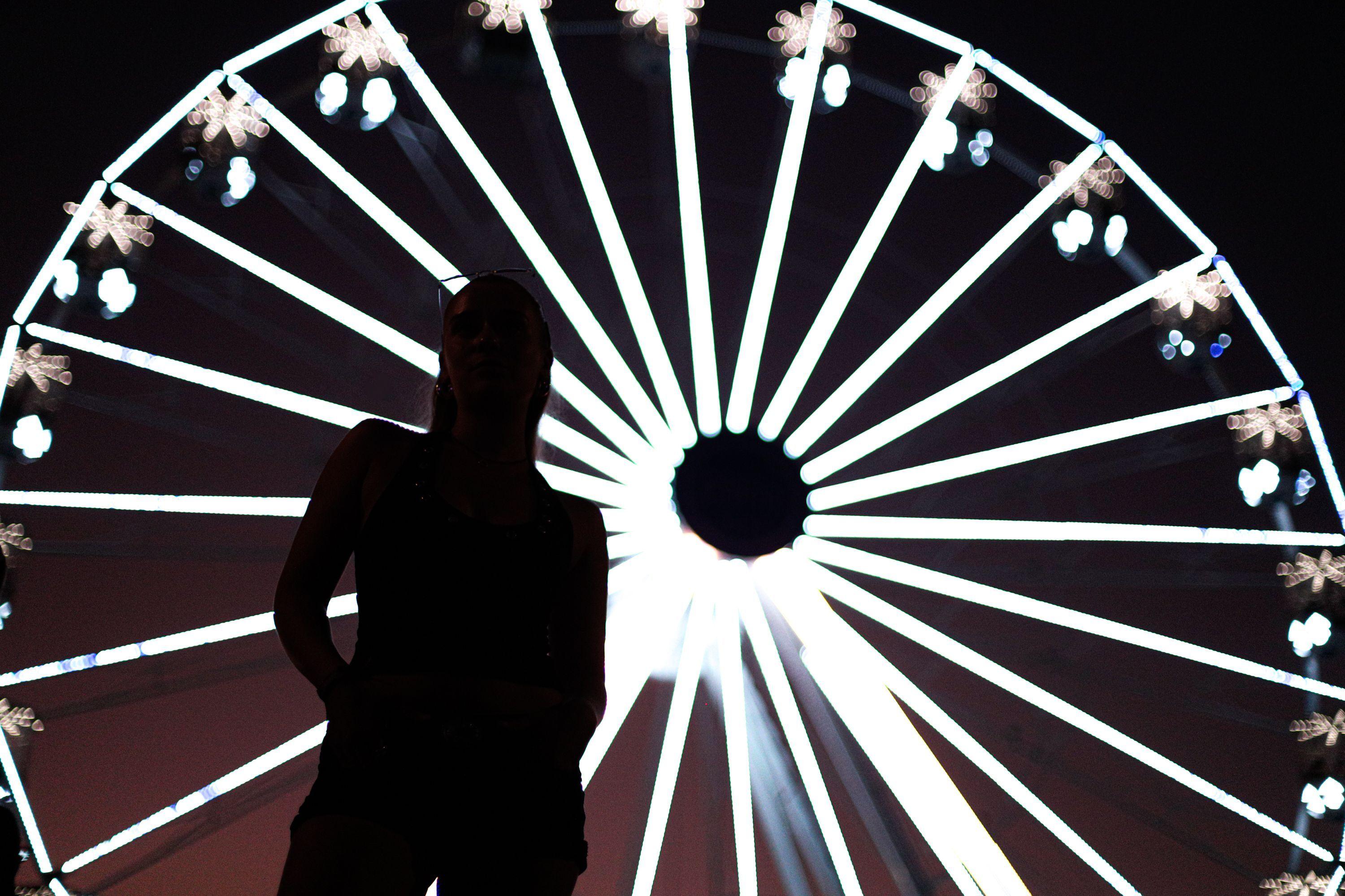 Silhueta de uma pessoa em primeiro plano, de p&eacute; e com as m&atilde;os atr&aacute;s do corpo, diante de uma grande roda-gigante iluminada &agrave; noite. As luzes brancas da roda formam linhas radiais brilhantes ao fundo, criando um efeito visual circular e contrastando com o corpo escuro da pessoa.