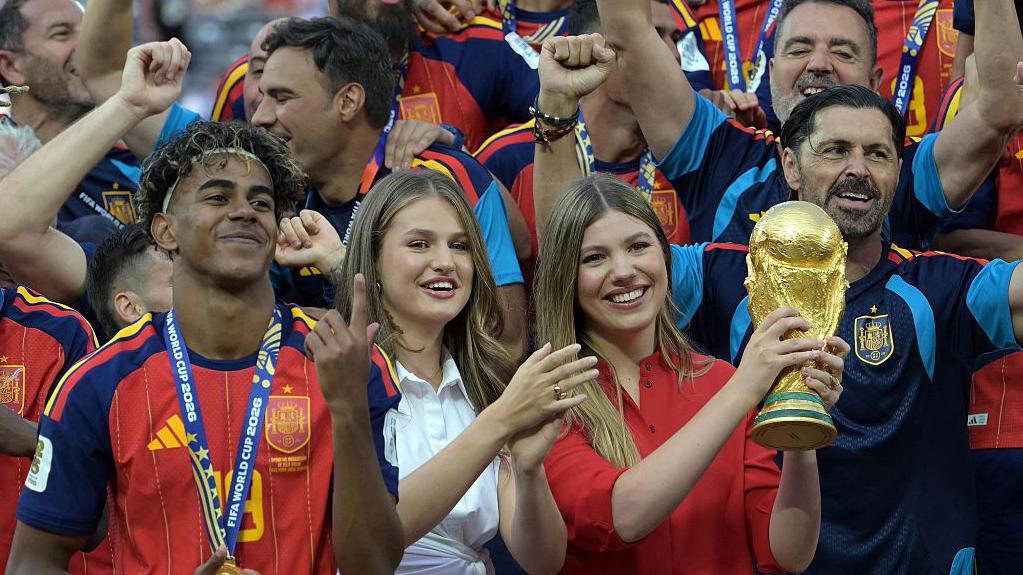 الأميرة صوفيا ترفع كأس العالم وسط احتفالات المنتخب الإسباني بالتتويج بلقب مونديال 2026، وإلى جانبها الأميرة ليونور والمهاجم لامين يامال، عقب الفوز على الأرجنتين في المباراة النهائية على ملعب نيويورك/نيوجيرسي في إيست راذرفورد بالولايات المتحدة، في 19 يوليو/تموز 2026.