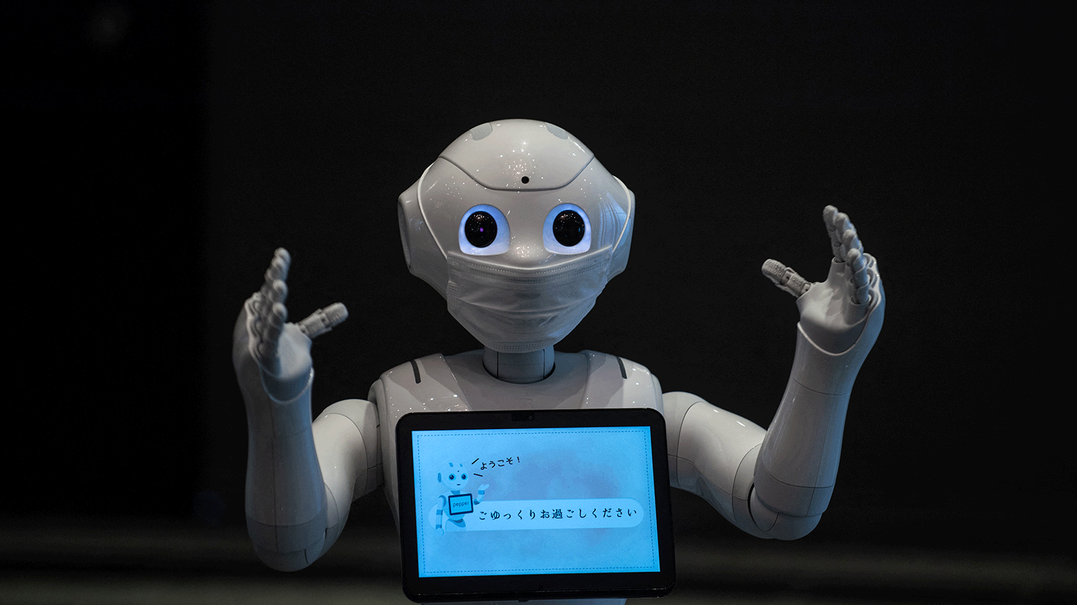 Um robô recepcionista, com os dois braços à frente, máscara no rosto e uma tela no peito.