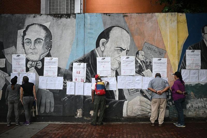 Local de votação na Venezuela em 25/05, com pessoas examinando papéis afixados a uma parede com pinturas