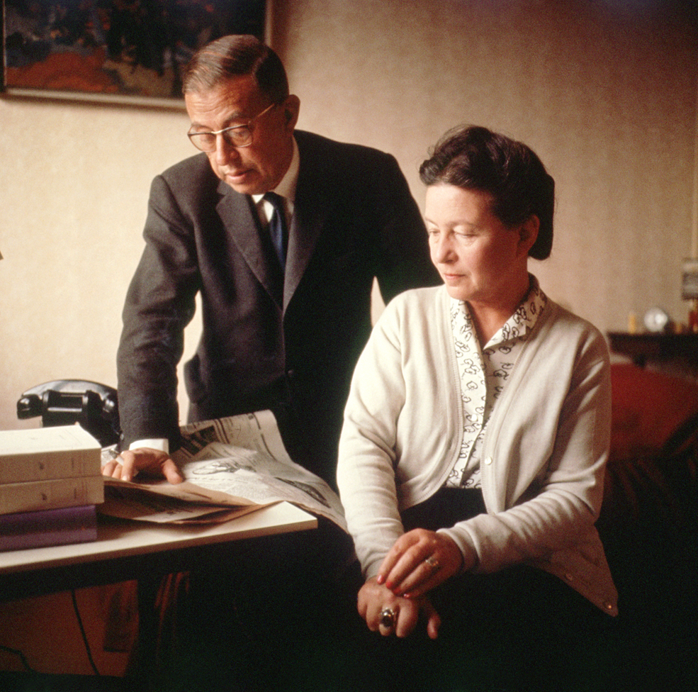 Sartre, parado, con un traje y corbata, apoya su mano en un periódico que está en un escritorio y al lado está De Beauvoir, sentada, con un saco beige y el pelo amarrado