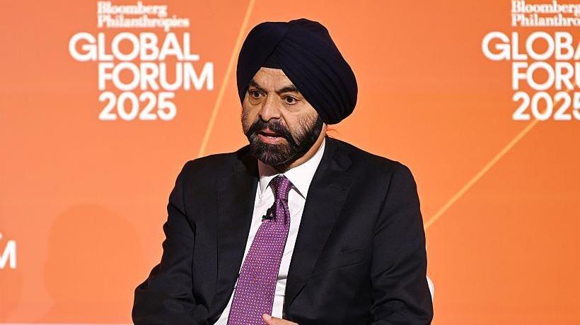 صورة لأجاي بانغا وهو يتحدث، مرتدياً عمامة سوداء وسترة سوداء وقميصاً أبيض وربطة عنق أرجوانية اللون، بها نقاط صغيرة بيضاء.