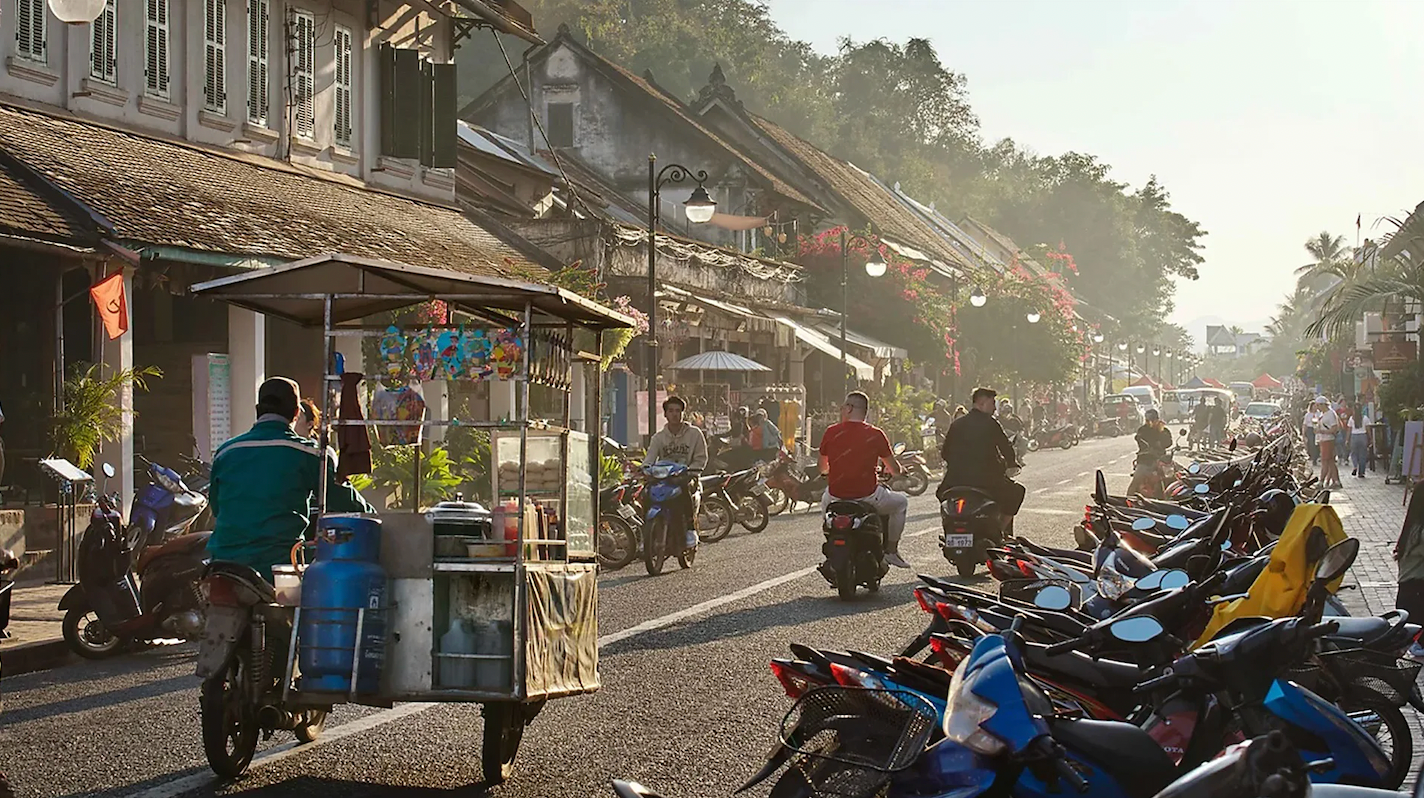 오토바이와 노점이 줄지어선 거리 위 몇 몇 사람들이 오토바이를 타고 거리를 지나간다. 