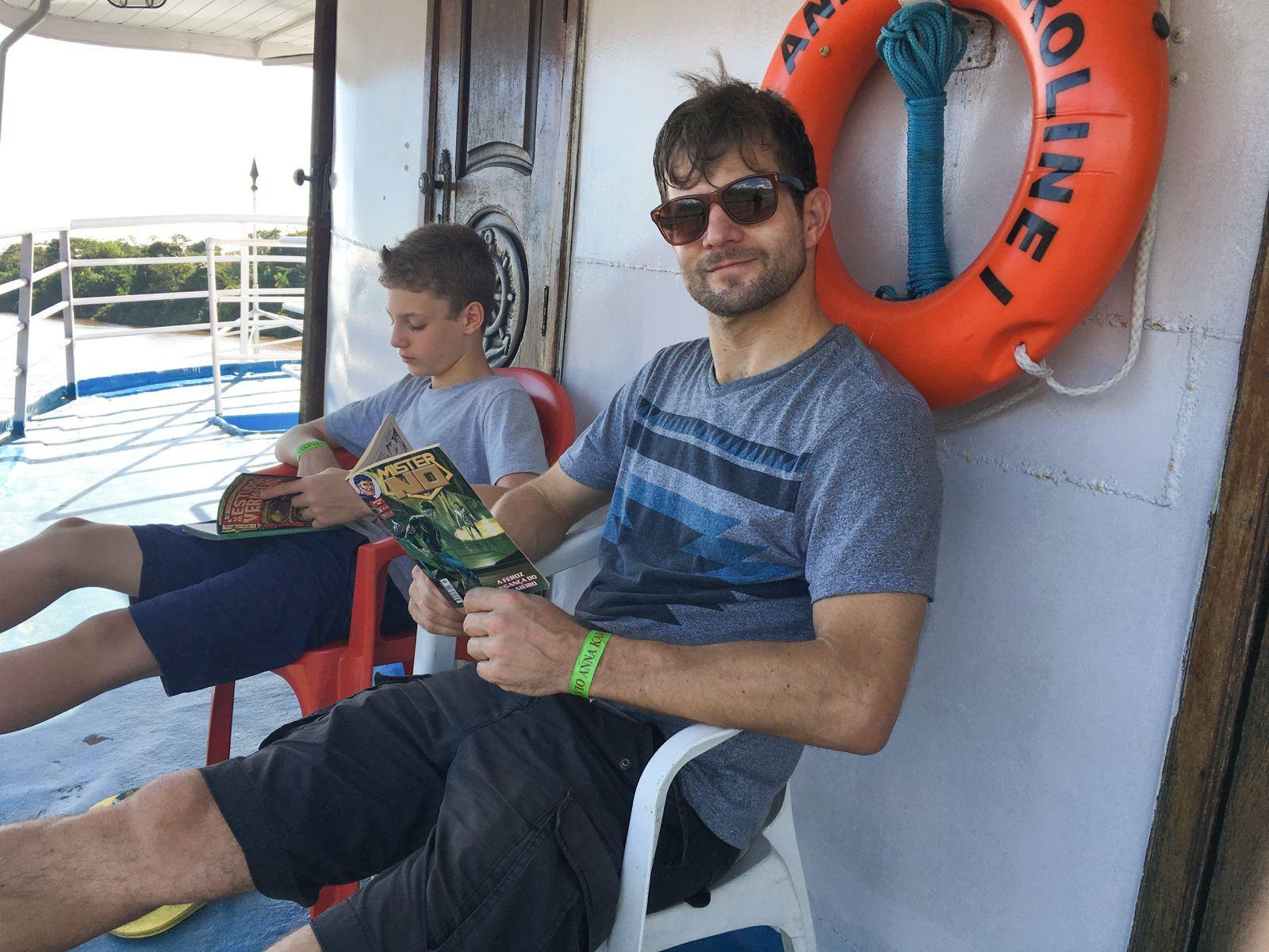 Um homem sentado em uma cadeira, dentro de um barco, lendo um gibi. Ao lado dele h&aacute; uma crian&ccedil;a.