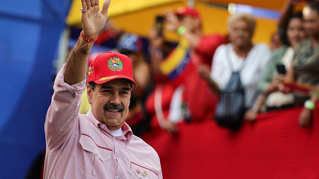 Nicolás maduro saluda con la mano extendida y una sonrisa a un grupo de personas. Tiene una gorra y camisa rojas. 