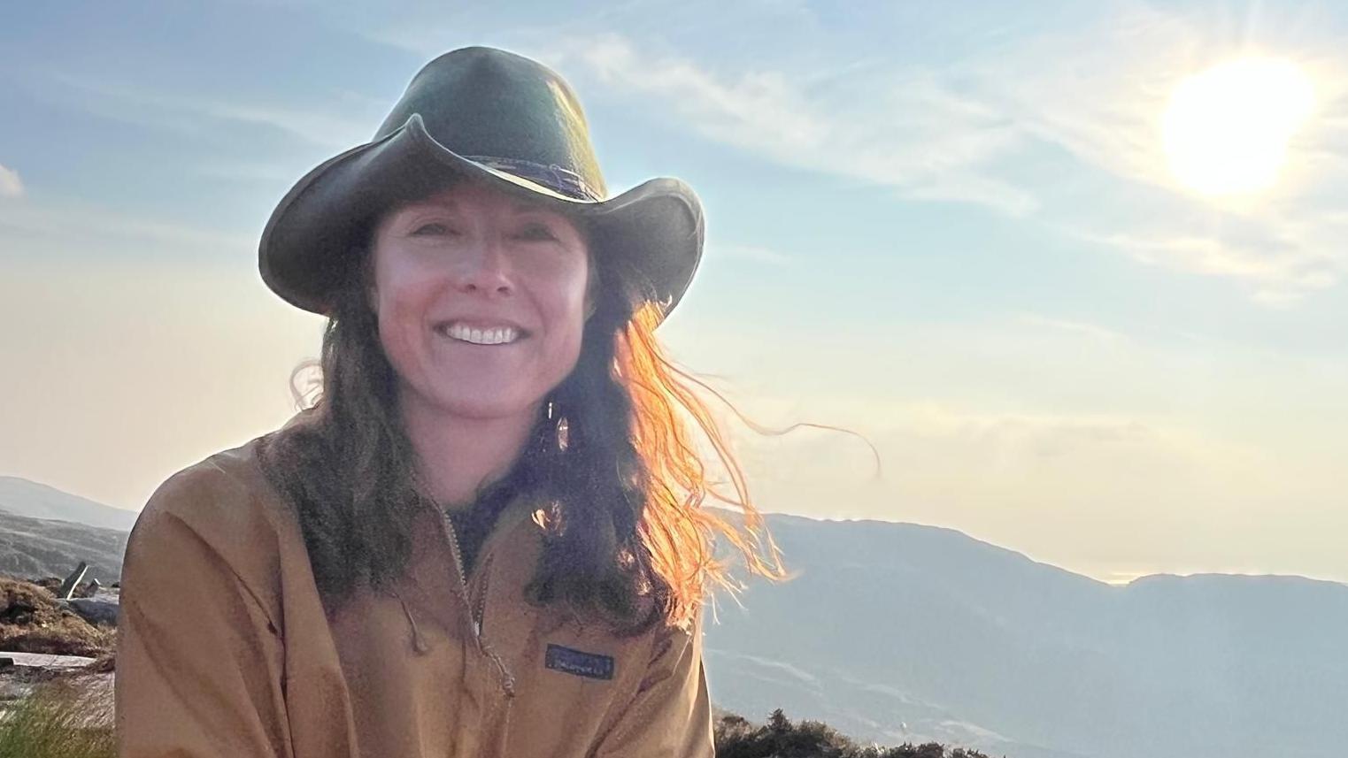 니나 라이언이 모자를 쓰고 카메라를 향해 미소 짓고 있으며, 뒤로는 산의 능선이 보인다