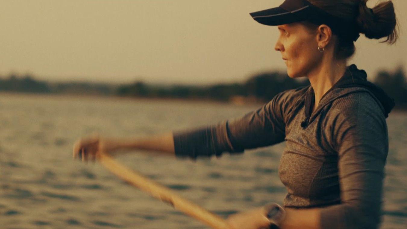 Uma mulher com viseira, brinco de argola e cabelo preso rema enquanto olha para o horizonte.