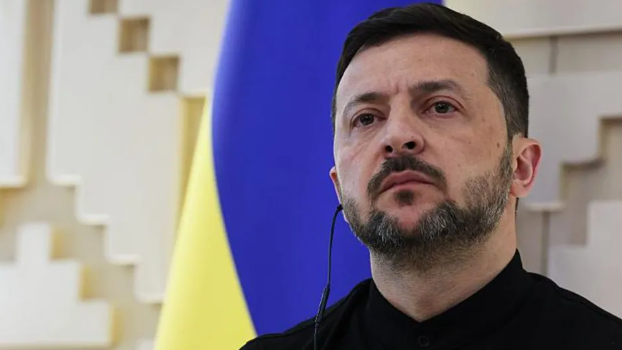 الرئيس الأوكراني فولوديمير زيلينسكي خلفه العلم الأوكراني وفي أذنه سماعة سوداء ويرتدي قميصاً أسود.