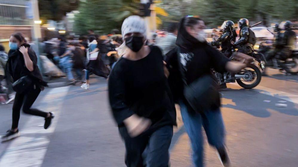 صورة ضبابية لسيدتين ترتديان كمامة، أثناء احتجاج بشوارع طهران، ويبد أنهم يركضون نحو الكاميرا، وبعض من المحتجين يهربون من شخصين يقودان دراجة نارية.