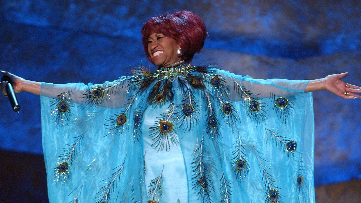 Celia Cruz, con vestido azul claro brillante, sonríe y abre los brazos mientras canta