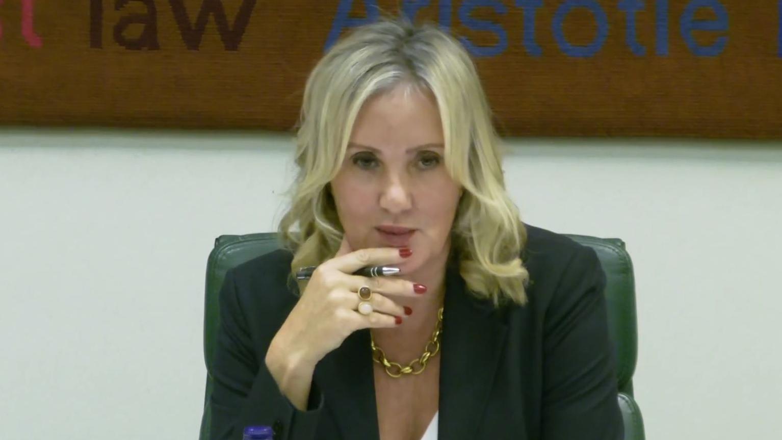 Caroline Dinenage - cabelo loiro, unhas vermelhas, segurando uma caneta, jaqueta escura, corrente dourada, sentada em uma cadeira.