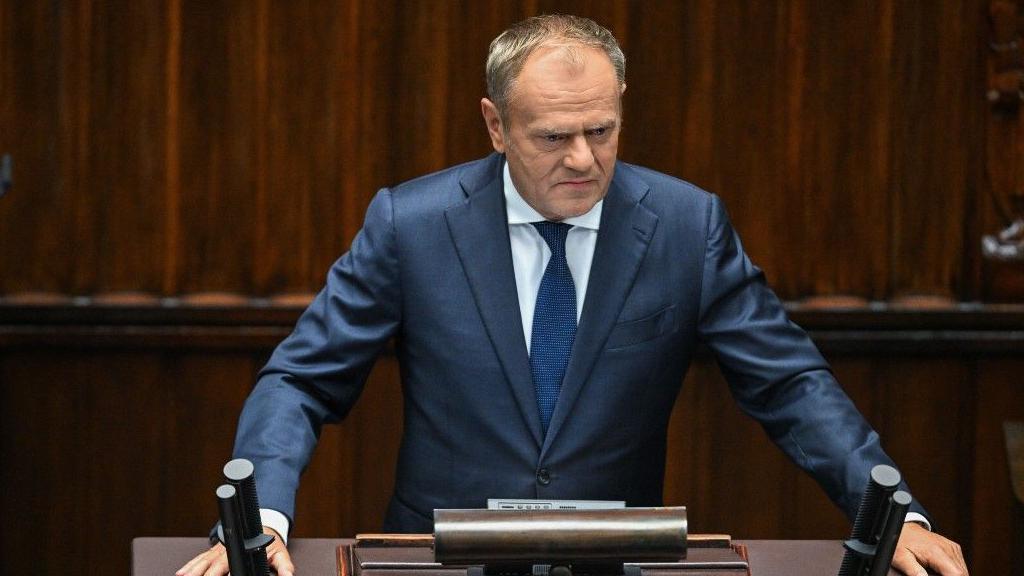 El primer ministro polaco, Donald Tusk, vestido con traje de chaqueta y gesto serio, hablando en el estrado del parlamento con sus manos extendidas sobre él. Todo el fondo y el estrado son de madera oscura. 