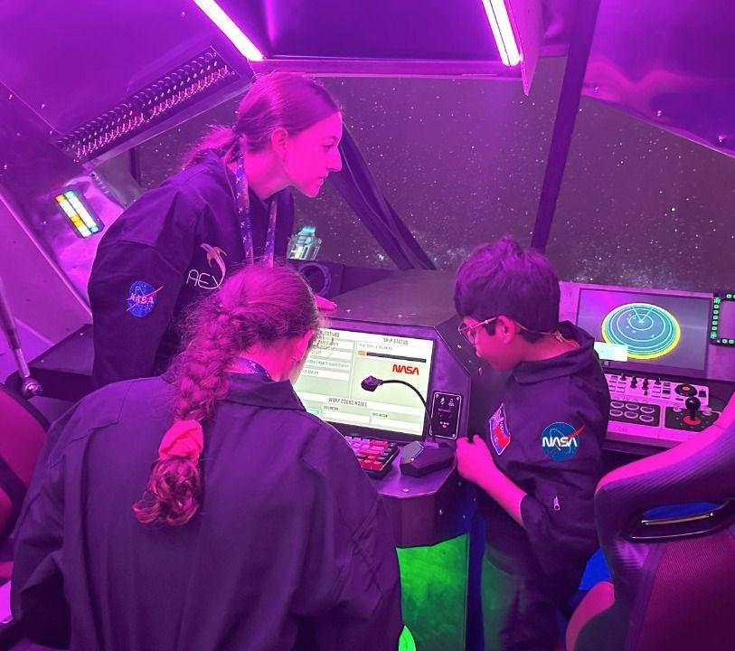 David Camacho, con otras dos mujeres jóvenes, todos vestidos con overoles de la NASA, están dentro de una cabina especial observando una computadora