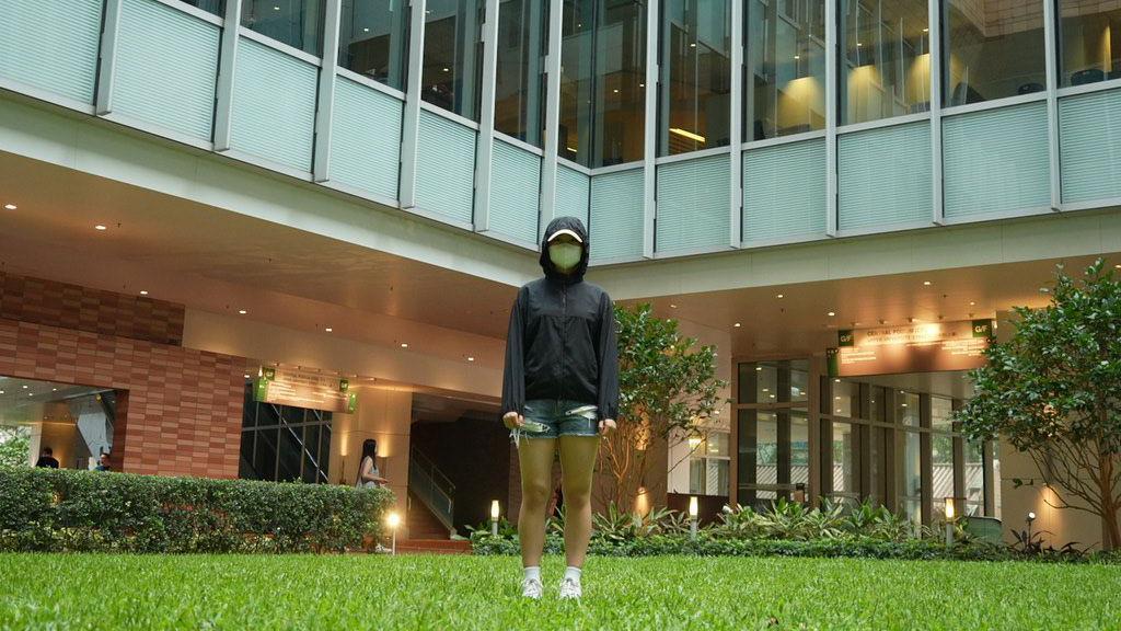 一名戴著帽子與口罩的女子站在大樓草坪前，背景是玻璃帷幕建築與燈光明亮的走廊。