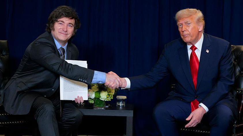Javier Milei vestido con traje negro y sentado en una silla le da la mano a Donald Trump, quien vestido de traje azul y corbata roja está sentado en una silla frente a él. Entre ambos hay una pequeña mesita con unas flores amarillas.