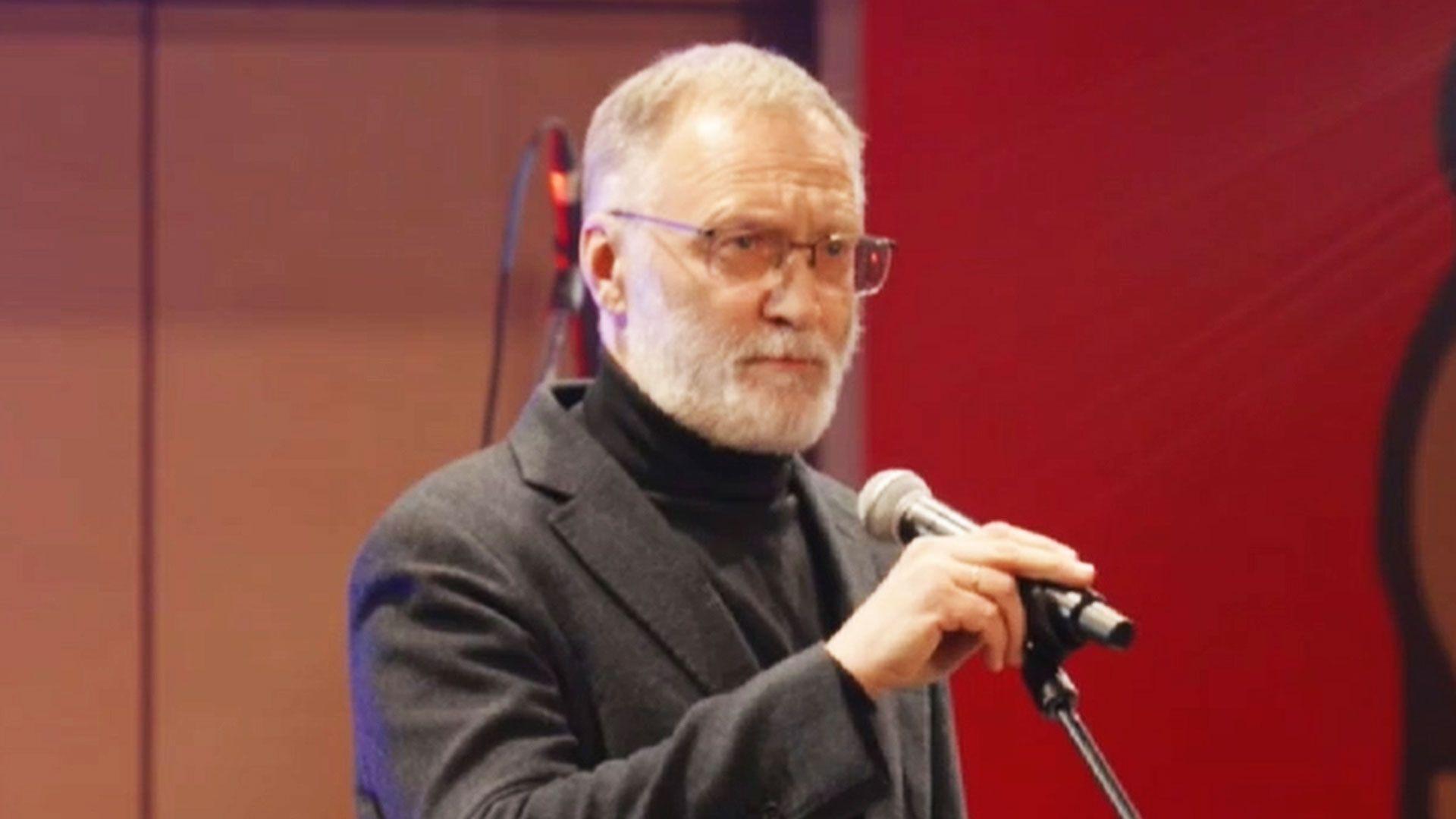 Sergei Mikheev veste terno cinza e gola alta preta. De &oacute;culos, com cabelos grisalhos curtos e barba rente ao rosto, ele aparece em um palco segurando um microfone