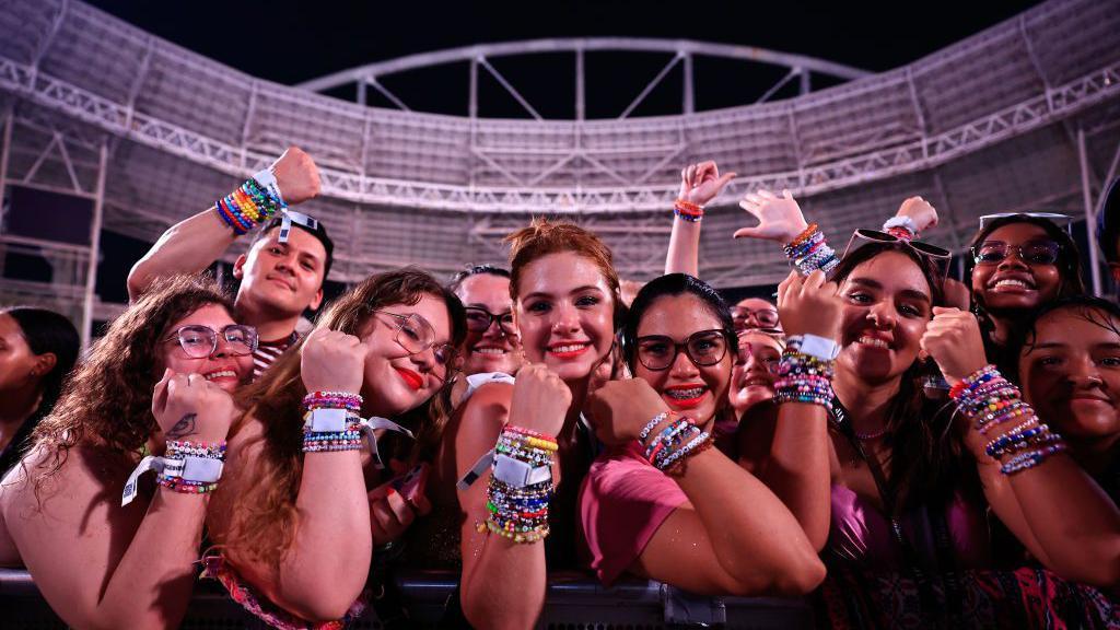 A imagem mostra um grupo de f&atilde;s de Taylor Swift posando na grade de um est&aacute;dio antes de um show, exibindo dezenas de pulseiras coloridas nos pulsos. Sorrindo para a c&acirc;mera, eles levantam os bra&ccedil;os para destacar os acess&oacute;rios, que se tornaram uma marca registrada entre admiradores da cantora, tendo ao fundo a estrutura iluminada da arena onde acontecer&aacute; a apresenta&ccedil;&atilde;o.