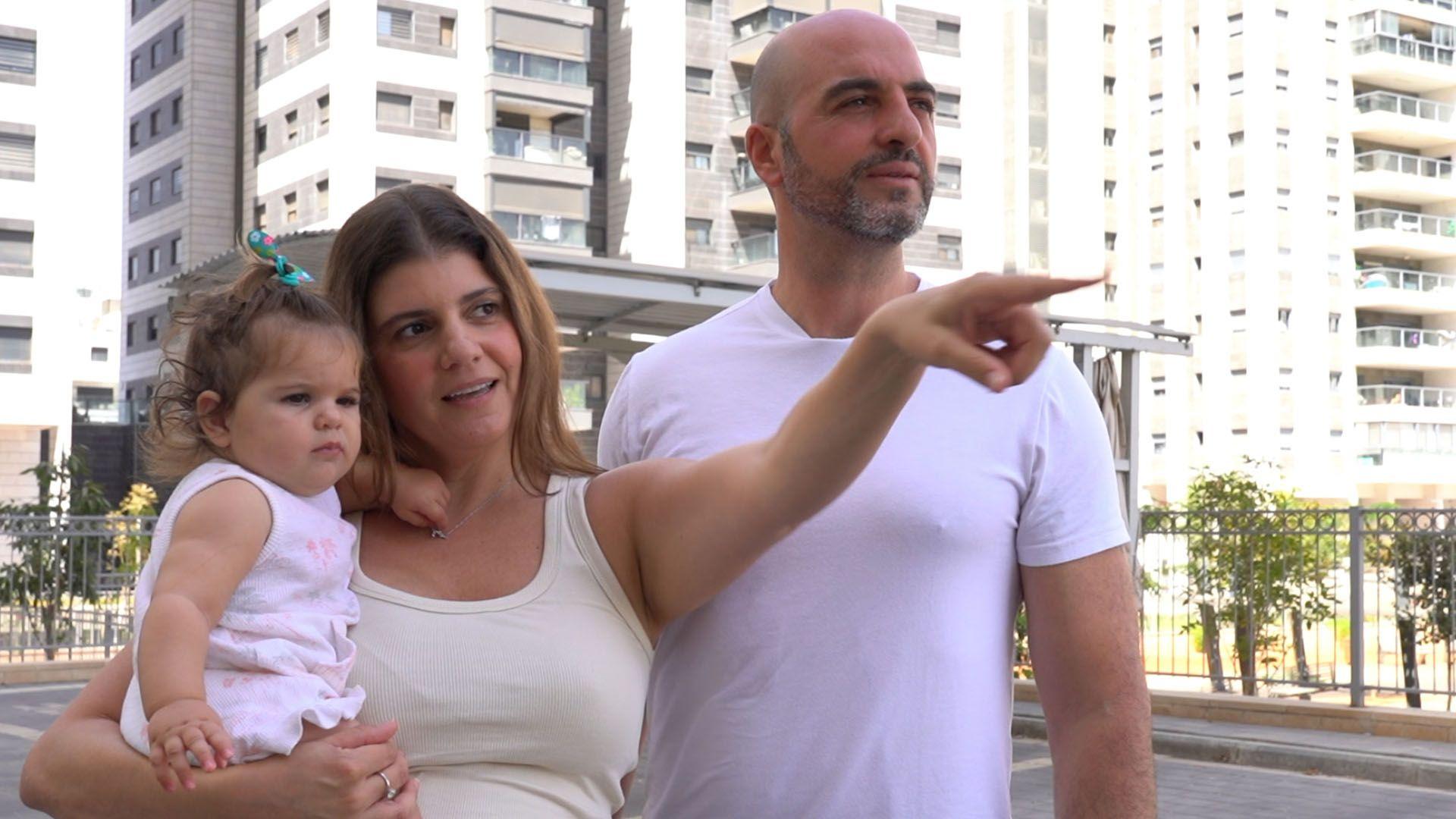 Una mujer sujeta a una niña pequeña con su brazo derecho y, con el izquierdo extendido, señala con el índice a un lugar al que mira la niña. Un hombre a su izquierda mira en la misma dirección. 