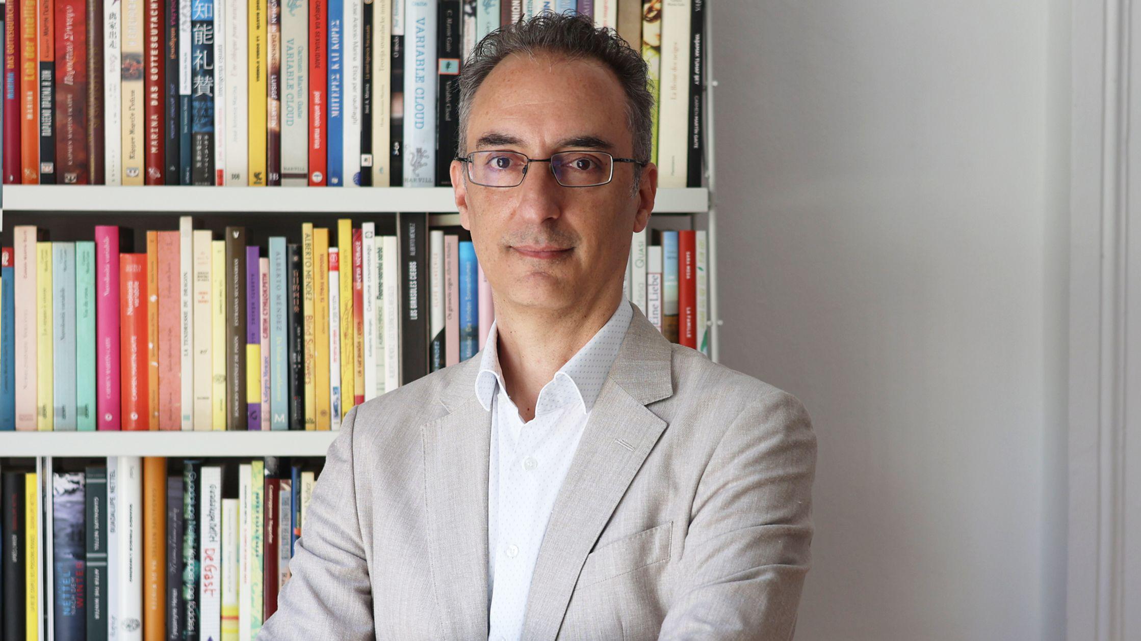 El analista y escritor Andrea Rizzi, con traje claro y gafas, delante de una estantería con libros