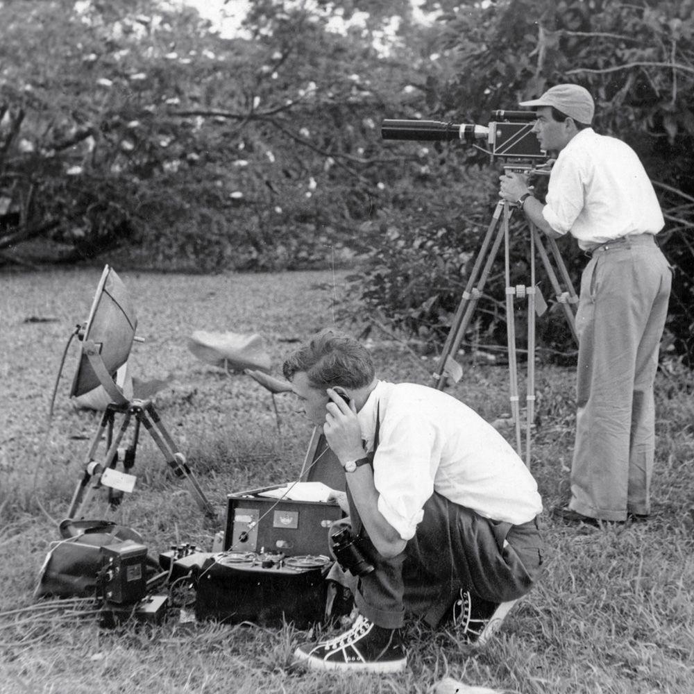Fotografía en blanco y negro de dos personas grabando en exteriores, con una cámara en trípode y equipo de audio entre la vegetación.