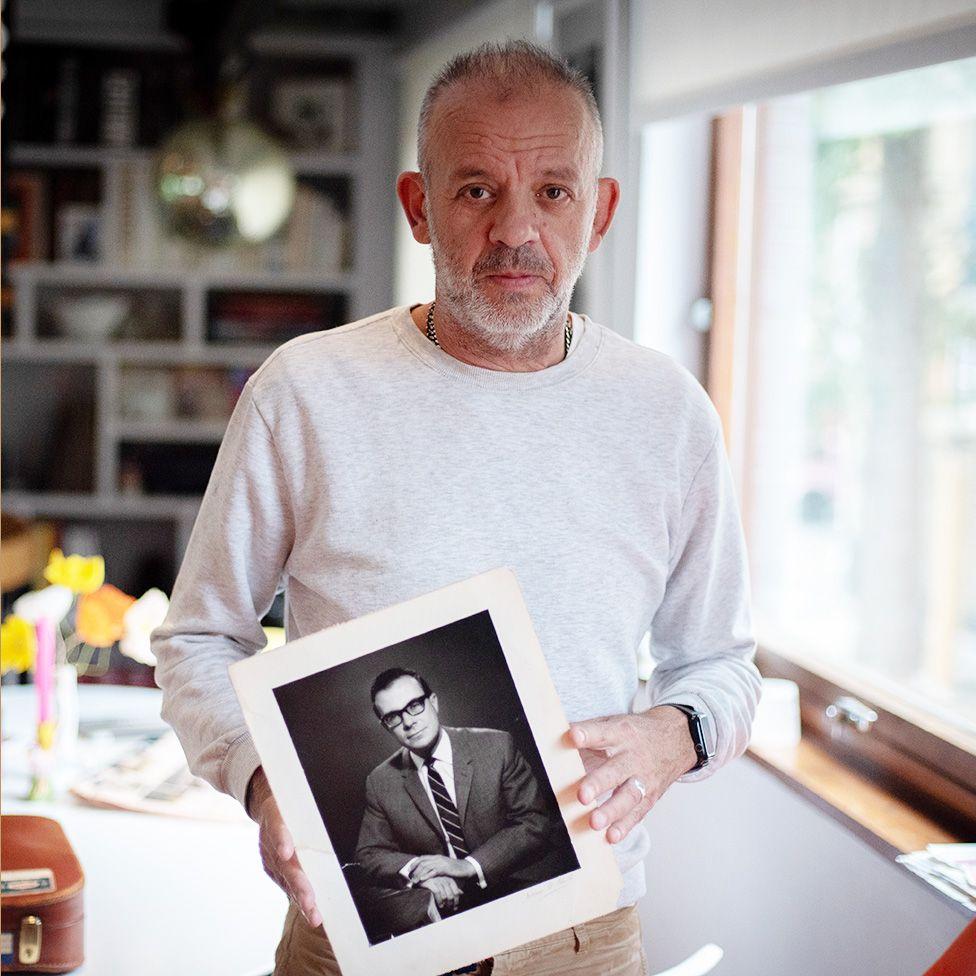 Antony Easton, vestindo uma camisa de manga comprida de cor clara, está em pé dentro de casa, segurando uma fotografia em preto e branco de seu pai, Peter Easton, vestido com terno e gravata. Atrás dele, há uma sala com estantes de livros e vários objetos, iluminada pela luz natural que entra por uma janela à direita. 
