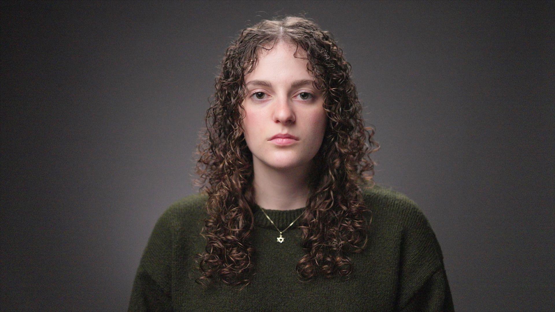 شابة تنظر إلى الكاميرا. لديها شعر طويل مجعد وترتدي سترة خضراء داكنة وسلسلة عليها نجمة داود حول رقبتها