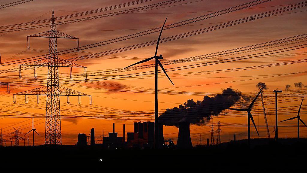 Esta fotografía, tomada al atardecer del 12 de noviembre de 2025, muestra aerogeneradores y postes de luz mientras se elevan los humos de las torres de refrigeración de la central termoeléctrica de lignito operada por el gigante energético alemán RWE, cerca de Neurath, en el oeste de Alemania. 