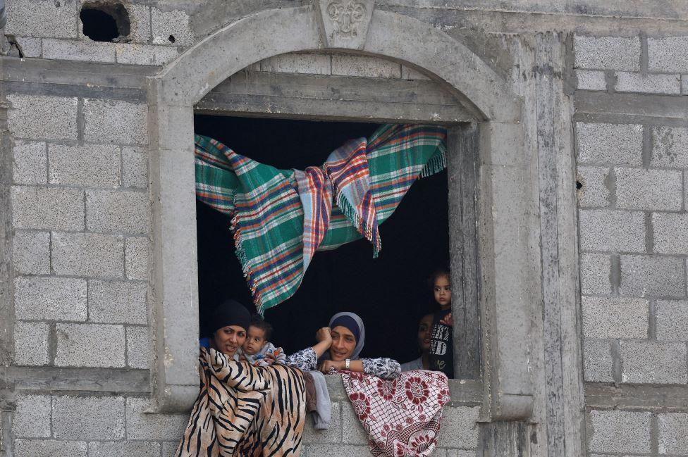 Mujeres y niños miran por la ventana de un edificio de piedra. Hay alfombras que cubren la ventana y cuelgan del techo.